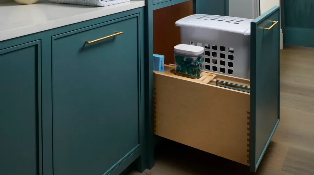 A concealed laundry hamper pulled out from deep green cabinetry